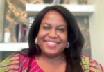 Portrait of a smiling woman with long dark hair wearing a pink and yellow patterned top, indoors with a blurred bookshelf in the background.