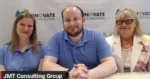 Three people sit side by side at a table in a conference room, wearing blue polo shirts, with a backdrop reading 'Innovate Consulting' and a bottom-left label 'JMT Consulting Group'.