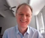 Smiling man in a light blue shirt on a video call, facing the camera in a bright office.