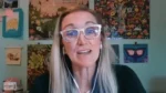 Woman with long gray hair and clear-framed glasses talking during a video call, wearing white earbuds, in a colorful poster-filled room.