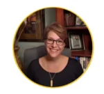 Portrait of a smiling woman with short light brown hair and glasses, seated in an office with bookshelves and framed pictures in the background.