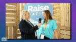 Two women talking at an interview, one holding a microphone, with a Raise banner in the background and wooden backdrop.